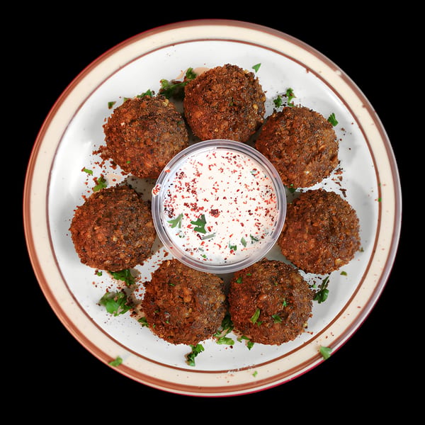 Six crispy falafel balls with a side of creamy white sauce, garnished with red spices and parsley.