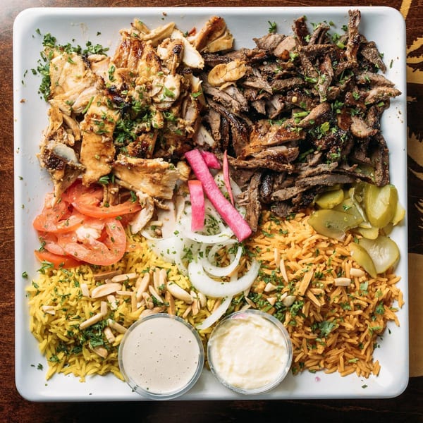 A large platter of Mediterranean food with chicken and beef shawarma, rice, tomatoes, onions, pickles, and sauces.